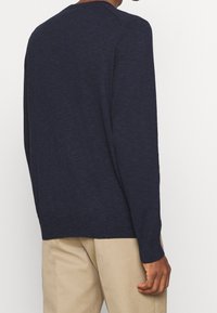 Man som bär en marinblå långärmad tröja och beige byxor, visad bakifrån mot en enkel vit bakgrund.