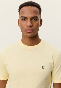 Κίτρινο βαμβακερό T-shirt με στρογγυλή λαιμόκοψη και κοντά μανίκια. Διαθέτει μικρό πράσινο κεντητό λογότυπο "II" στο αριστερό μέρος του στήθους. Λείο υφή.