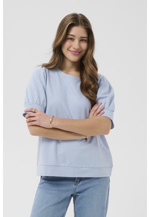 Tânără cu păr lung ondulat, purtând un top bleu cu mâneci bufante și blugi bleu deschis, zâmbind cu brațele încrucișate.