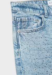 Jeans de mezclilla azul claro con un acabado desgastado, que presentan tachuelas metálicas dispersas en la parte delantera y un diseño clásico de cinco bolsillos.