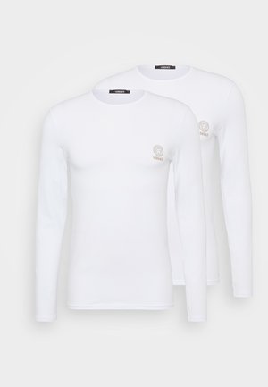 Langärmlige weiße T-Shirts aus Baumwolle mit rundem Halsausschnitt, die ein kleines goldenes Löwenlogo auf der oberen linken Brust zeigen. Zwei identische Shirts.