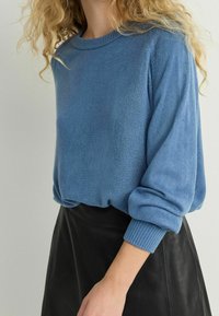 Niebieski sweter dzianinowy z żebrowanym okrągłym dekoltem i mankietami. Lekko luźny krój z teksturowanym materiałem. Noszony na czarnej skórzanej spódnicy.