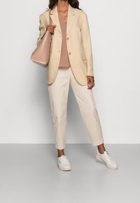 Blazer beige con rever tagliati, top marrone chiaro, pantaloni crema e sneakers bianche con dettagli verdi. Tiene una borsa a spalla rosa pallido.