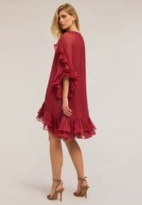 Žena v průhledných, červených šatech s volány ke kolenům a hnědých vysokých podpatcích, stojící s otočeným zády a hledící přes rameno.