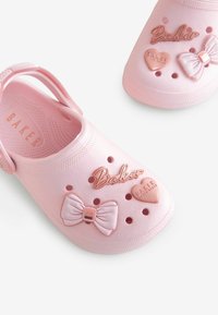 Roze rubberen klompen hebben reliëfontwerpen van harten en strikken, met ventilatiegaten en een achterband voor een veilige pasvorm. "Baker" logo weergegeven.