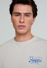 Light gray short-sleeve t-shirt features a blue graphic logo on the left chest reading "Scalpers." The fabric appears soft with a smooth texture.