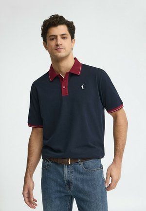 Man die een navyblauw poloshirt draagt met een rode kraag en mouwboord, blauwe spijkerbroek en een bruine riem, staand tegen een effen grijze achtergrond.