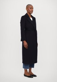 Dlouhý tmavě námořnický kabát s páskem v pase, klopami s výřezem a bočními kapsami, v kombinaci s krátkými džínami a černými balerínami. Hladká textura.