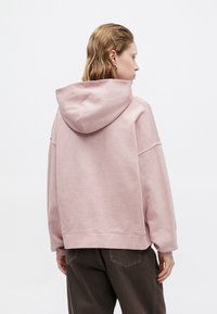 Rózsaszín, laza szabású kapucnis felső, széles ujjaival és húzózsinórral, puha anyagból készült. Részletei között szerepel a bordázott mandzsetta és alsó szegély, barna nadrággal párosítva.