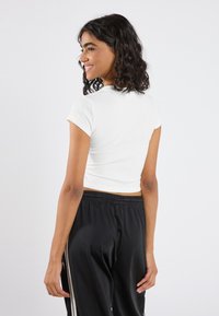 Fehér bordázott rövidujjú crop top fekete sportnadrággal, amelynek húzózsinóros derékrésze és oldalsó csíkjai vannak, hátulról nézve.