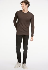 Brauner langärmeliger Strickpullover mit Rundhalsausschnitt, kombiniert mit schwarzen Slim-Fit-Jeans und weißen Turnschuhen. Der Pullover hat ein dezentes Logodetail.