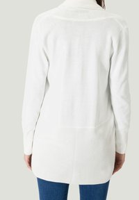 Cardigan tricoté blanc avec un col montant, des manches longues et un dos texturé. L'ourlet est plus long à l'arrière, porté avec un jean bleu.