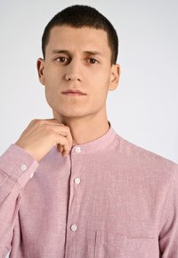 Camicia di lino rosa con colletto a fascia, tasca frontale e bottoni bianchi. La mano del modello è appoggiata vicino al colletto, con uno sfondo neutro.