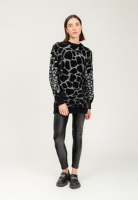 Donna che indossa un lungo maglione a strisce nero e grigio, leggings in pelle nera e mocassini neri con dettagli in catena dorata, in piedi su uno sfondo neutro.