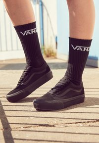 Baskets basses en toile noire avec des accents en daim, dotées d'un design à lacets et d'une semelle en caoutchouc ton sur ton, associées à des chaussettes noires côtelées affichant le logo Vans.