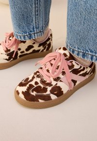 Braune und weiße Kuhflecken-Sneaker mit rosa Schnürsenkeln, kombiniert mit beigen Socken und blauen Jeans.