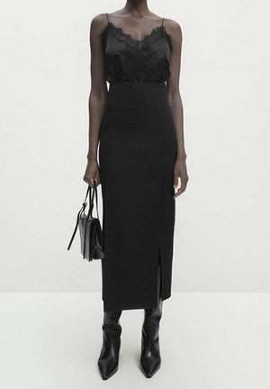 Woman wearing a black lace-trimmed camisole, high-waist long skirt with front slit, black pointed boots, carrying a black handbag.