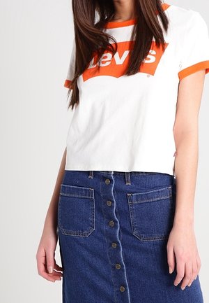 Mujer vistiendo una camiseta corta blanca de Levi's con ribete y logotipo naranja, combinada con una falda de mezclilla con botones en la parte delantera y grandes bolsillos frontales.
