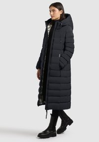 Lange zwarte gewatteerde jas met capuchon, gestikt design, ritssluiting over de hele lengte en glanzende zilveren voering. Gecombineerd met zwarte enkellaarzen.