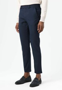 Marineblauwe, op maat gemaakte broek van een gladde stof, met een strakke pasvorm, zijzakken en een knoopsluiting. Gecombineerd met donkerbruine suède instapschoenen.