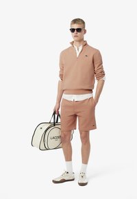 Sudadera de cuello con cremallera de color rosa claro, pantalones cortos a juego, camiseta blanca debajo y gafas de sol negras. Sosteniendo un bolso de deporte de Lacoste beige con detalles en negro.