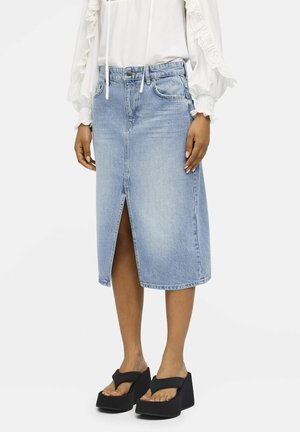 Kvinde iført lys blå knælængde denimnederdel med frontslids, hvid bluse med blonde detaljer og sorte platformsandaler.