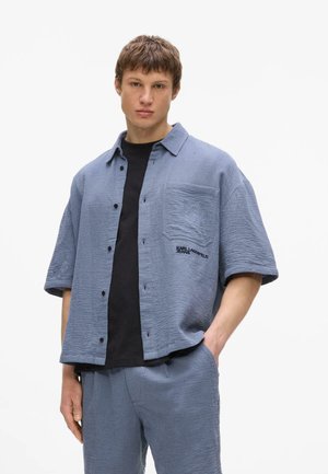 Joven con camisa azul de manga corta texturizada y pantalones a juego con camiseta negra debajo, de pie con una mano en el bolsillo.