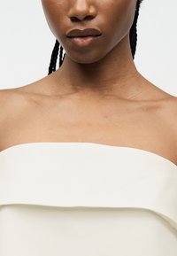 Κοντινό πλάνο από το κάτω μέρος του προσώπου, το λαιμό και τους ώμους ατόμου που φοράει λευκό τοπ χωρίς λουριά, με φόντο απλό ανοιχτόχρωμο.