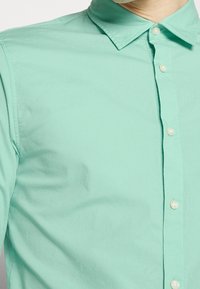 Camicia button-up verde chiaro con colletto classico, caratterizzata da una texture liscia e bottoni bianchi lungo la patta davanti.