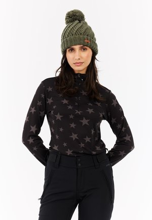 Schwarzes Langarmshirt mit Sternmuster, mit Viertelreißverschluss; kombiniert mit enganliegenden schwarzen Hosen. Grüne gestrickte Mütze mit Pompon-Detail.