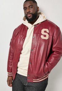 Man draagt een crèmekleurige hoodie onder een rode leren varsityjas met een beige patch in de vorm van een "S", staand tegen een effen lichte achtergrond.