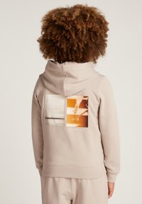 Bambino con capelli ricci che indossa una felpa beige e pantaloni. Sul retro della felpa ci sono due stampe grafiche e il testo "Calvin Klein Jeans".