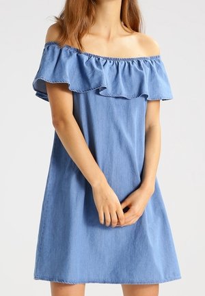 Frau trägt ein hellblaues schulterfreies Kleid mit geschnürtem Ausschnitt, Hände sanft vor dem Körper gefaltet, neutraler Hintergrund.