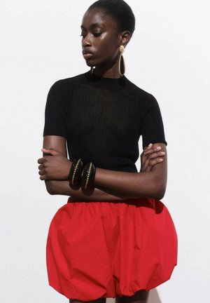 Zwarte geribbelde top met korte mouwen, gecombineerd met losse rode shorts. Accessoires omvatten grote gouden oorbellen en stevige zwarte armbanden.