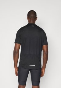 Schwarzes kurzärmliges Radtrikot aus strukturiertem Stoff, mit einer Rückentasche und reflektierenden Akzenten. Getragen über schwarzen Radshorts.