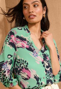 Daugiakampė marška, žalia, su abstrakčiais raštais rožinės, juodos ir mėlynos spalvų. Mygtukų užsegimas su plonybėmis rankovių kraštais.