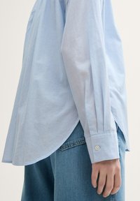 Camisa de manga larga azul claro en tejido suave con puños abotonados y aberturas laterales, combinada con pantalones cortos de denim de cintura alta.