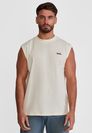 Mann med kort brunt hår og skjegg, iført ermeløs off-white skjorte med liten svart logo og blå jeans, stående mot en ensfarget bakgrunn.