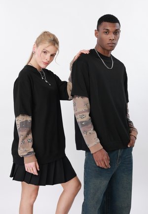 Due persone stanno fianco a fianco indossando maglie nere oversize sovrapposte a maniche lunghe beige mimetiche; una indossa una gonna, l'altra un paio di jeans.