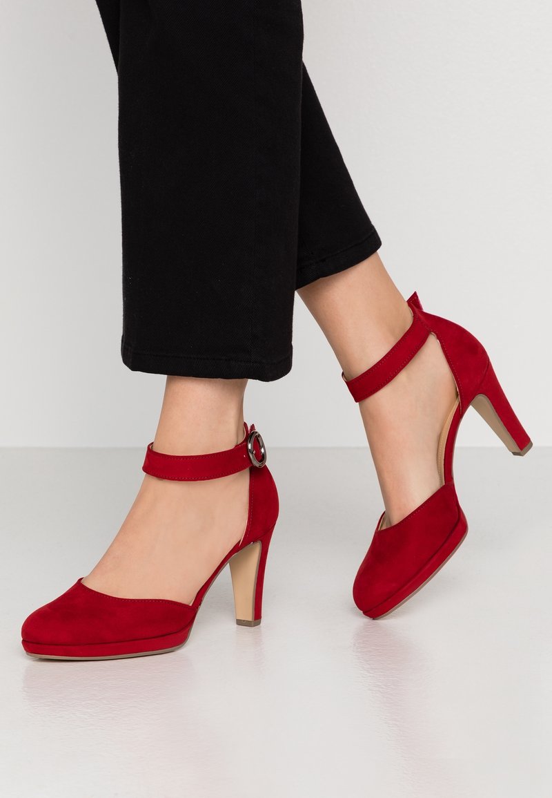 Pieds portant des chaussures à talons hauts en suède rouge avec des brides à la cheville et des boucles argentées, associées à un pantalon noir coupé au-dessus de la cheville sur un fond uni.