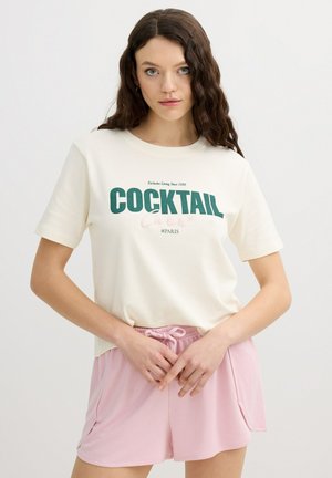 Mujer con el pelo largo y oscuro que lleva una camiseta blanca con el texto "COCKTAIL #PARIS" y pantalones cortos rosas con cordón, de pie frente a un fondo liso.