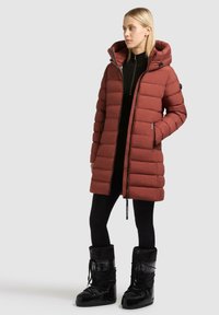 Lungo giubbotto imbottito bordeaux con cappuccio, caratterizzato da una trapuntatura orizzontale, una zip frontale e tasche laterali; abbinato a stivaletti neri da neve.