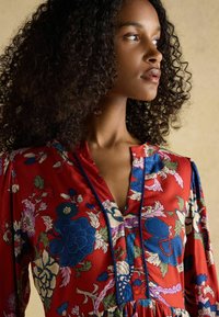 Rotes Blumenkleid mit blauen und grünen Mustern, V-Ausschnitt und filigranen Details. Aus einem glatten, leichten Stoff gefertigt.