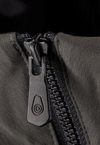 Sort lynlåsskovl på grå stof, med en tekstureret trækstrop og synlige tænder. Nærbillede viser stofvæv og syningsdetaljer.