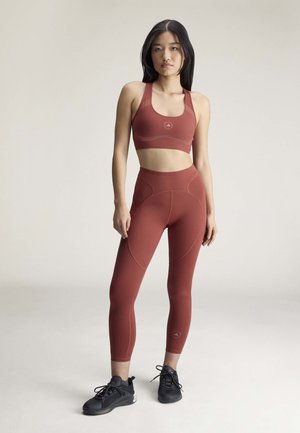 Mlada žena stoji u sportskom grudnjaku i tajicama boje hrđe s crnim tenisicama, na jednobojnoj pozadini.