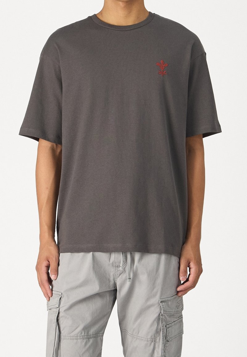 Homme portant un t-shirt à manches courtes gris foncé avec un petit emblème floral rouge sur le côté gauche de la poitrine et un pantalon cargo gris clair.