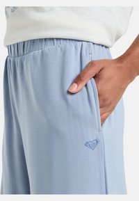 Pantalones de chándal azul claro con una cintura elástica, bolsillos laterales y un pequeño logo de corazón bordado en el lado izquierdo. Textura de tela suave.