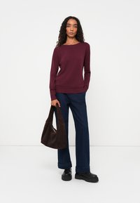 Pull en tricot bordeaux avec un col rond, associé à un pantalon large bleu foncé. Le mannequin tient un sac à main en suède marron foncé. Chaussures à plateforme noires.