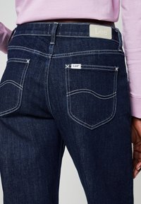 Vaqueros de mezclilla azul oscuro con costuras blancas en contraste, que cuentan con una etiqueta de cuero en la cintura y un pequeño logo en el bolsillo trasero.