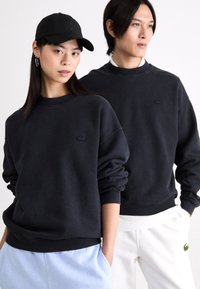 Két ember sötét tengerészkék, kerek nyakú, bordás mandzsettával ellátott pulóvert visel. A pulóvereken a Lacoste logó található a mellrészen.